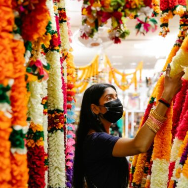 A woman wearing a mask shops for vibrant handmade floral garlands indoors.