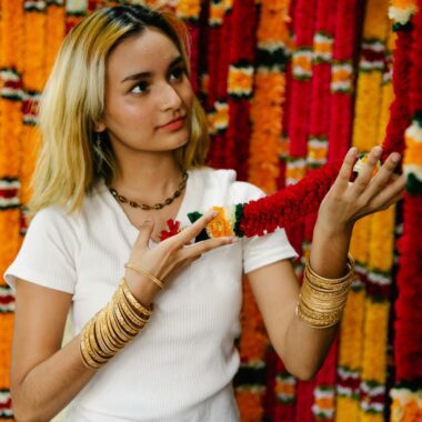 A woman with blonde hair and bracelets holds a vibrant floral garland indoors.