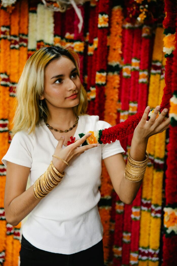 A woman with blonde hair and bracelets holds a vibrant floral garland indoors.